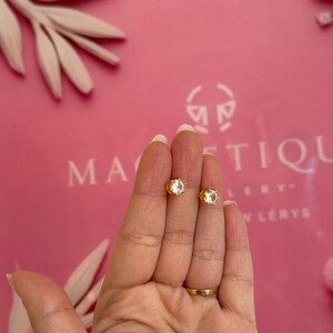 May include: A pair of gold-colored stud earrings with clear, faceted gemstones. The earrings are held in a hand against a pink background with the word "MAGNIFIQUE" visible. The earrings are small and elegant.