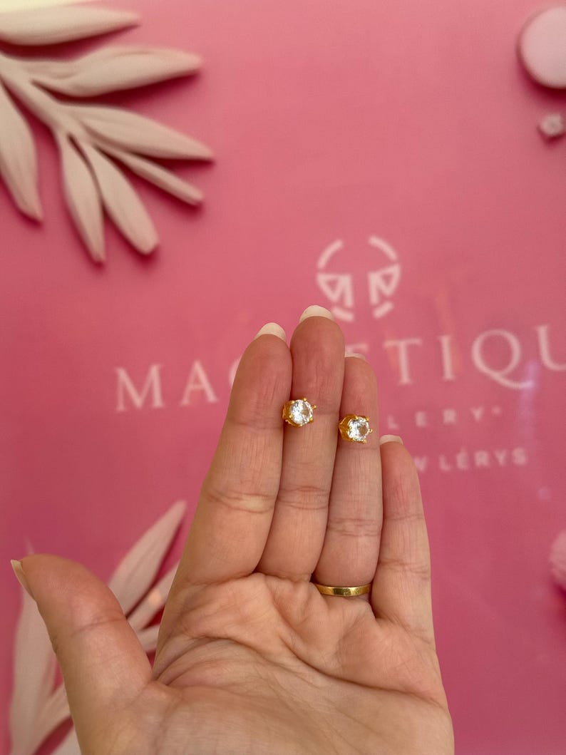 May include: A pair of gold-colored stud earrings with clear, faceted gemstones. The earrings are held in a hand against a pink background with the word "MAGNIFIQUE" visible. The earrings are small and elegant.