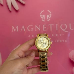 May include: Gold-toned wristwatch with a round face and a crystal-studded bezel. The watch has a gold band and a white watch face with three smaller dials. The background is pink with the word "MAGNETIQUE" printed on it.
