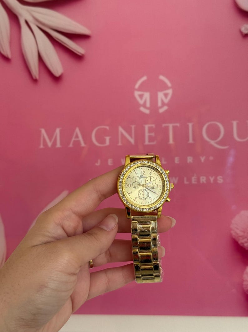 May include: Gold-toned wristwatch with a round face and a crystal-studded bezel. The watch has a gold band and a white watch face with three smaller dials. The background is pink with the word "MAGNETIQUE" printed on it.
