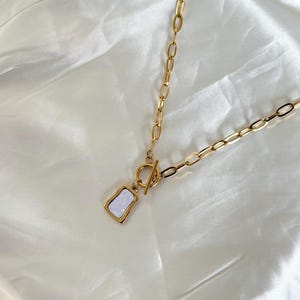 May include: Gold-coloured necklace with a rectangular pendant featuring a white, iridescent inlay. The necklace has a toggle clasp and a chain with alternating oval and elongated links. The jewellery is displayed on a white, satin-like fabric.