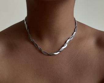 May include: A silver-colored necklace with a braided design. The necklace is worn around the neck and sits close to the skin. The necklace is made of a shiny metal and has a smooth texture. The jewelry is a choker style.