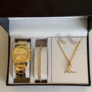 May include: A gold-toned watch, bracelet, necklace, earrings, and ring set displayed in a black box with a white interior. The watch and jewelry are embellished with clear crystals. The watch has a gold face and band.