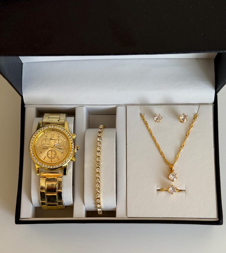 May include: A gold-toned watch, bracelet, necklace, earrings, and ring set displayed in a black box with a white interior. The watch and jewelry are embellished with clear crystals. The watch has a gold face and band.