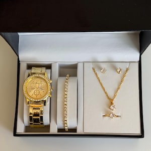 May include: A gold-toned jewelry set in a black box. The set includes a watch, bracelet, necklace, earrings, and a ring. The watch and bracelet have a row of clear stones. The necklace and ring have a single clear stone.