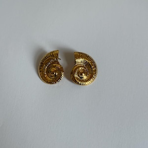 May include: A pair of gold-coloured earrings. Each earring is shaped like a spiral, with a textured surface. The earrings are set against a white background. The earrings are likely made of metal.