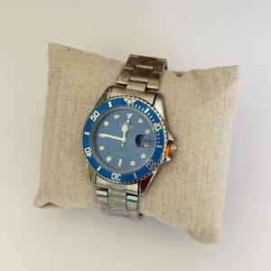 May include: A silver-tone analogue wristwatch with a blue dial and a matching blue rotating bezel. The watch features a stainless steel link bracelet, luminous hour markers, a date window at the three o'clock position, and the text YANKO QUARTZ.