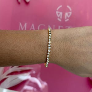 May include: A gold tennis bracelet with clear, sparkling stones. The bracelet is worn on a wrist, showcasing the delicate design and the brilliance of the stones. The background is a vibrant pink with the word "MAGNET" visible.