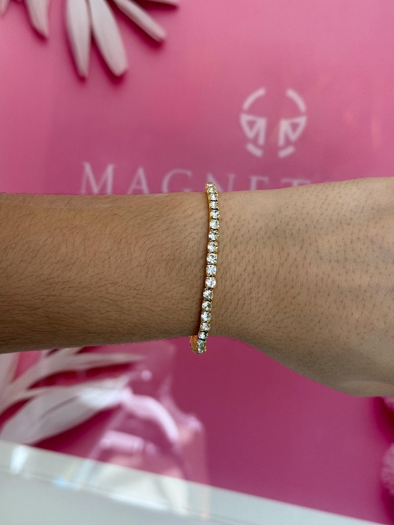 May include: A gold tennis bracelet with clear, sparkling stones. The bracelet is worn on a wrist, showcasing the delicate design and the brilliance of the stones. The background is a vibrant pink with the word "MAGNET" visible.