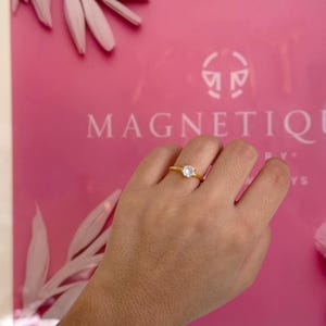 May include: A gold-colored ring with a clear, round gemstone. The ring is set on a hand against a pink background with the word "MAGNETIQUE" visible. The ring's band is thin and simple.