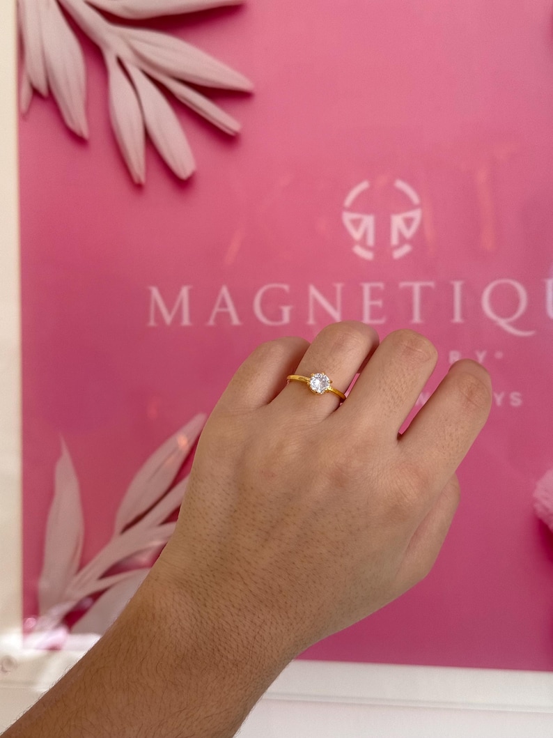 May include: A gold-colored ring with a clear, round gemstone. The ring is set on a hand against a pink background with the word "MAGNETIQUE" visible. The ring's band is thin and simple.