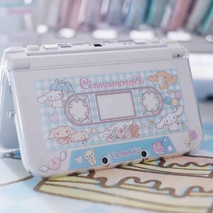 May include: White handheld gaming console with a Cinnamoroll design. The design includes a cassette tape illustration, hearts, and other cute graphics. The console is open, revealing the screen and buttons. The text "Cinnamoroll" is visible.