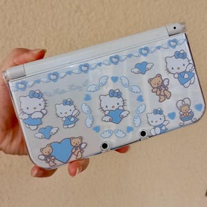 Puede incluir: Una consola de juegos portátil Nintendo 3DS blanca con una funda protectora transparente. La funda presenta un patrón repetido de personajes de Hello Kitty, corazones azules y ositos de peluche. El texto "Hello Kitty" también es visible.