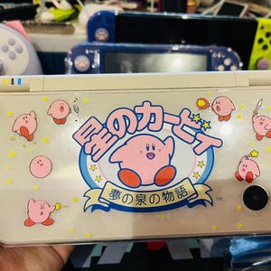 May include: A handheld gaming console with a cream-coloured case and a colourful Kirby design. The design includes the character Kirby, stars, and Japanese text. The console has a camera and a power button.