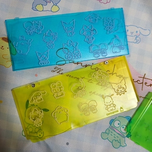 Puede incluir: Dos estuches de plástico transparente, uno azul y otro amarillo, con diseños en relieve de personajes de dibujos animados. El estuche azul presenta personajes como Hello Kitty. El estuche amarillo tiene otros personajes.