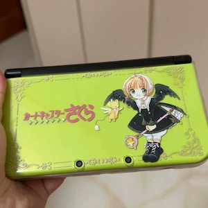 Puede incluir: Consola de juegos portátil Nintendo 3DS de color verde lima con una ilustración de dibujos animados de una niña con alas y texto japonés. La consola tiene una parte superior negra y adornos dorados.