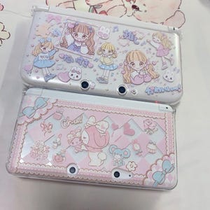 May include: Two white Nintendo 3DS consoles with decorative covers. The top console features cartoon girls and Japanese text. The bottom console has a pink and white design with bows, hearts, and the word "Chu!"