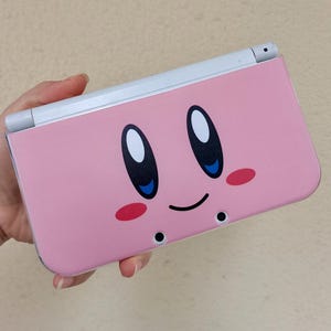 May include: A pink handheld games console with a cartoon face design. The face has large black and white eyes with blue accents, rosy cheeks, and a smiling mouth. The console has a white hinge and is held in a hand.