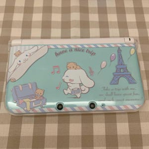 May include: A light blue Nintendo 3DS case featuring cartoon characters and travel-themed illustrations. The case has the text "have a nice trip" and "Take a trip with me, we shall have great fun, and such sweet memories!"