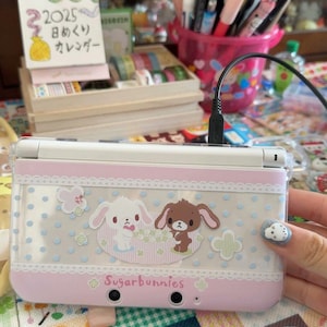 May include: A pink and white Nintendo 3DS handheld game console with a decorative Sugarbunnies design. The design features cartoon bunnies, polka dots, and lace trim. The console is connected to a charging cable.