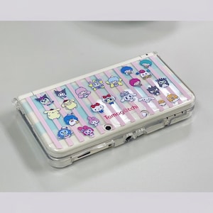 May include: A white handheld gaming console with a clear protective case. The case is decorated with pink and blue striped pattern and cartoon characters. The text "Tamag@tchi" is printed on the case.