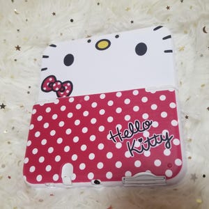 May include: A white and red Hello Kitty-themed handheld gaming device case. The top section is white with black eyes, a yellow nose, and a red bow. The bottom is red with white polka dots and the words "Hello Kitty".