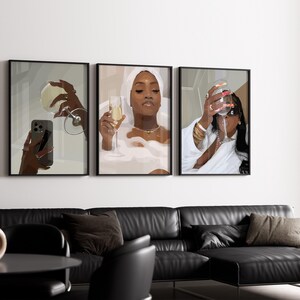 May include: Three framed art prints depicting women enjoying champagne. One print shows a woman in a bathtub, another shows a woman taking a selfie with a glass, and the third shows a woman drinking champagne. The prints are displayed above a black leather sofa.