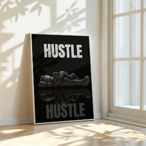 May include: Black and white framed art print featuring the word "HUSTLE" in bold white letters. A black figure is lying down, with a reflection below. The artwork is displayed against a white wall, next to a window.