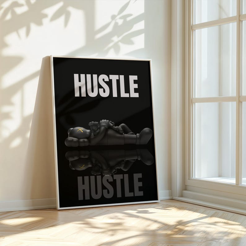 May include: Black and white framed art print featuring the word "HUSTLE" in bold white letters. A black figure is lying down, with a reflection below. The artwork is displayed against a white wall, next to a window.