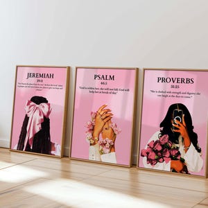 May include: Three framed art prints with pink backgrounds and gold frames. Each print features a woman and text from the Bible: Jeremiah 29:11, Psalm 46:5, and Proverbs 31:25. The art style is modern and inspirational.