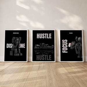 May include: Three framed black and white art prints. The prints feature stylized black figures with white text that reads "DISCIPLINE", "HUSTLE", and "FOCUS". The figures are in various poses, and the prints are displayed on a wooden floor.