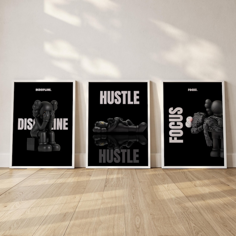 May include: Three framed black and white art prints. The prints feature stylized black figures with white text that reads "DISCIPLINE", "HUSTLE", and "FOCUS". The figures are in various poses, and the prints are displayed on a wooden floor.