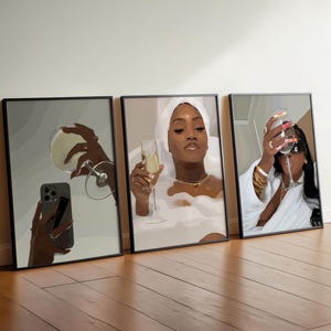 May include: Three framed art prints leaning against a wall. Each print features a woman with champagne. One holds a phone, another is in a bathtub, and the third is wearing sunglasses. The art has a modern, minimalist style.