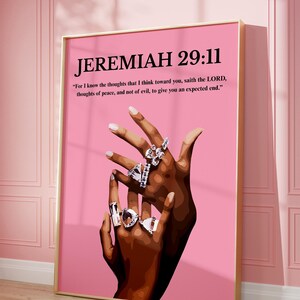 May include: A framed print with the text "JEREMIAH 29:11" and a quote on a pink background. The artwork features a pair of hands adorned with multiple silver rings. The frame is gold.