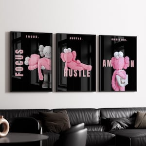 May include: Three framed art pieces with black backgrounds and pink text that read "FOCUS", "HUSTLE", and "AMBITION". Each frame features a different plush toy character in a unique pose. The art is displayed above a black leather sofa.