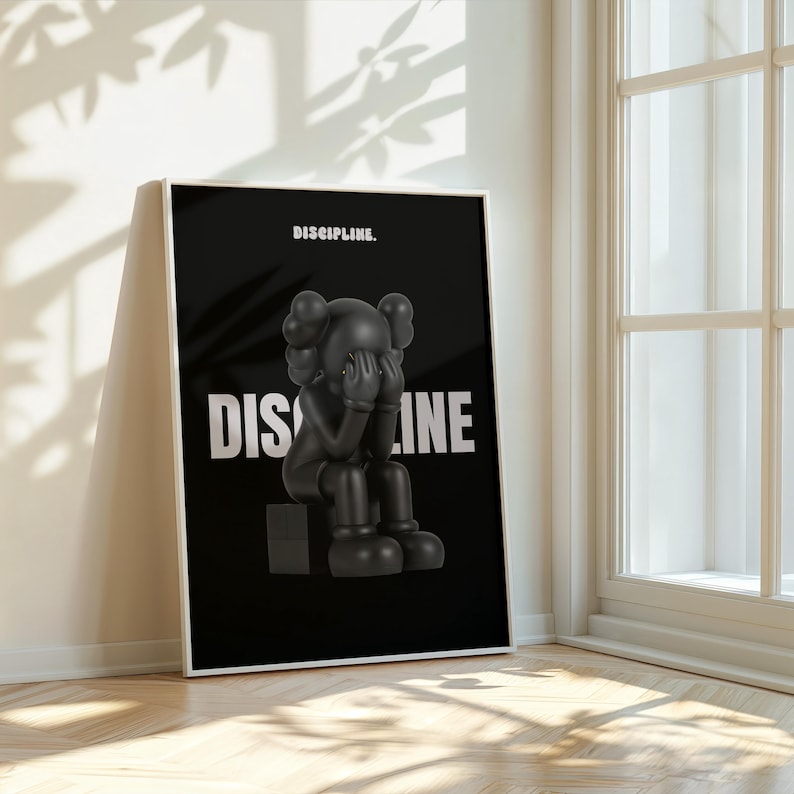 May include: Black framed art print featuring a black figure with hands covering its face, sitting on a black cube. The word "DISCIPLINE" is printed in white, with the word "DIS" on the left and "INE" on the right. The word "DISCIPLINE" is printed above the figure.