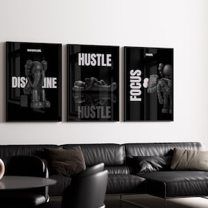May include: Three framed black and white art prints with the words "DISCIPLINE", "HUSTLE", and "FOCUS" in white. The prints feature stylized figures. The art hangs above a black leather sectional sofa in a modern living room setting.