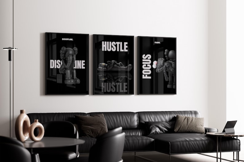 May include: Three framed black and white art prints with the words "DISCIPLINE", "HUSTLE", and "FOCUS" in white. The prints feature stylized figures. The art hangs above a black leather sectional sofa in a modern living room setting.