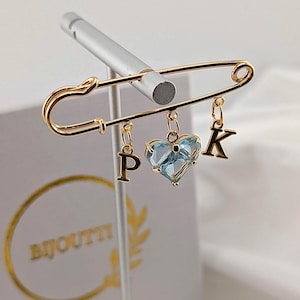 Op de afbeelding: Een goudkleurige veiligheidsspeld-broche met drie bungelende bedels: een letter P, een lichtblauwe hartvormige gefacetteerde edelsteen en een letter K. De metalen speld heeft een gepolijste afwerking en de bedels zijn bevestigd met kleine ringetjes.
