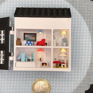 Könnte beinhalten: Ein Miniatur-Puppenhaus mit schwarzem Dach, weißen Wänden und vier Zimmern. Die Zimmer sind mit winzigen Möbeln eingerichtet, darunter ein roter Stuhl, blaue Stühle und ein Tisch. Eine Ein-Euro-Münze befindet sich neben dem Puppenhaus.