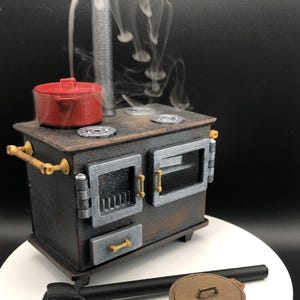 Op de afbeelding: Een miniatuur houtgestookte kachel met een rode pot erop en een bruine pot op de vloer. De kachel heeft een zilveren deur en een schoorsteen waar rook uit komt.