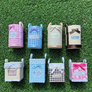 Puede incluir: Ocho cestas de lavandería en miniatura con varios colores y estampados, incluyendo cuadros vichy rosas, cuadros vichy azules, beige y flores. Cada cesta tiene un pequeño lazo, dos asas y una etiqueta con la palabra Laundry en el frente.