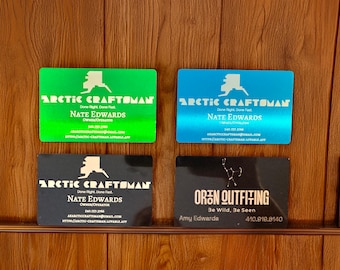 May include: Four business cards are displayed against a wooden background. Two cards are green and blue, featuring the text "ARCTIC CRAFTSMAN" and "Nate Edwards." The other two cards are black, with the same "ARCTIC CRAFTSMAN" and "Nate Edwards" text, and "OREN OUTFITING" with "Amy Edwards."