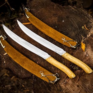May include: Three silver blades with wooden handles and gold accents are displayed on a weathered wooden surface. Each blade is sheathed in a brown leather scabbard with gold clasps. The blades feature etched designs.