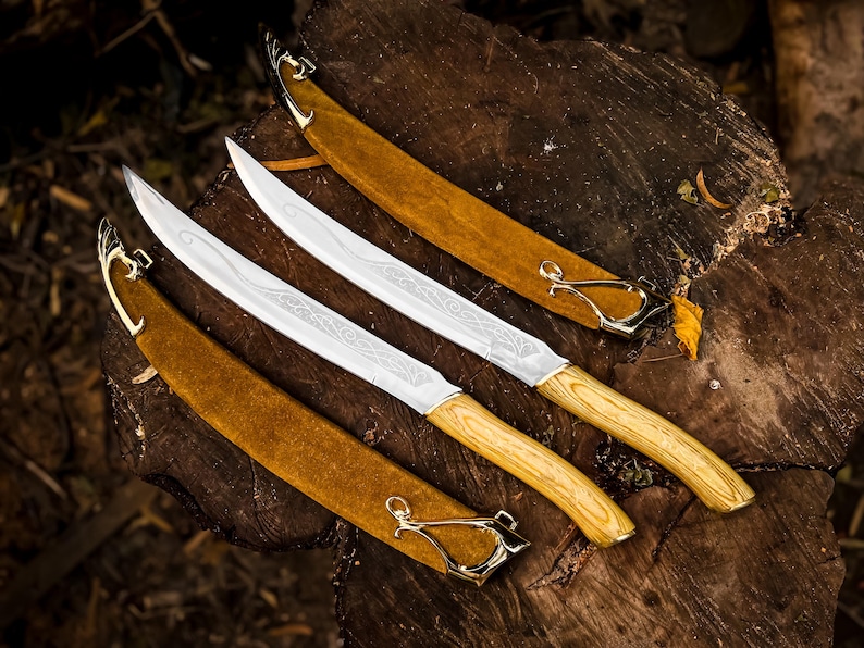 May include: Three silver blades with wooden handles and gold accents are displayed on a weathered wooden surface. Each blade is sheathed in a brown leather scabbard with gold clasps. The blades feature etched designs.