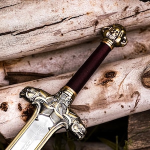 May include: A detailed close-up of a decorative sword with a silver blade and ornate gold-colored hilt. The handle is wrapped in a deep burgundy material. The sword rests on a background of stacked, light-colored logs.