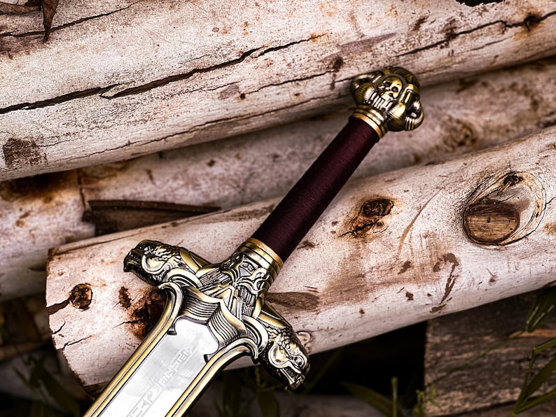 May include: A detailed close-up of a decorative sword with a silver blade and ornate gold-colored hilt. The handle is wrapped in a deep burgundy material. The sword rests on a background of stacked, light-colored logs.