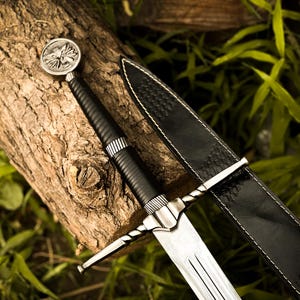 May include: A medieval-style sword with a silver blade and a black handle, resting on a weathered log. The sword has a decorative silver pommel and a black leather sheath. The background features green foliage.