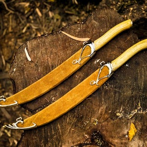 May include: Two decorative curved knives with light brown suede-like sheaths and light wood handles. The blades have silver-coloured metal accents. The knives are resting on a dark brown, textured wooden surface.