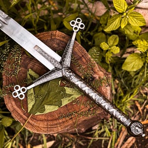 May include: A detailed, silver-colored sword with an ornate handle and blade rests on a wooden surface. The sword's handle features intricate designs and a decorative pommel. The background includes green foliage and a wooden stump.